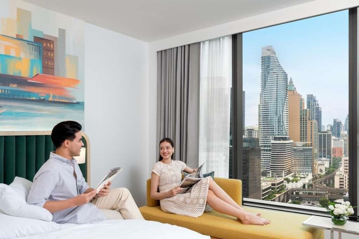 A hotel room with a bed, a sofa, a large window with a city view, and a painting on the wall.
