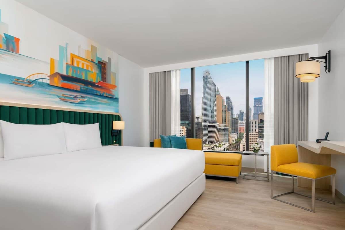 A hotel room with a large bed, a desk, a chair, and a cityscape view through large windows.