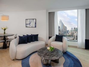 A modern living room with a sofa, armchairs, a round coffee table, and a city view through large windows.