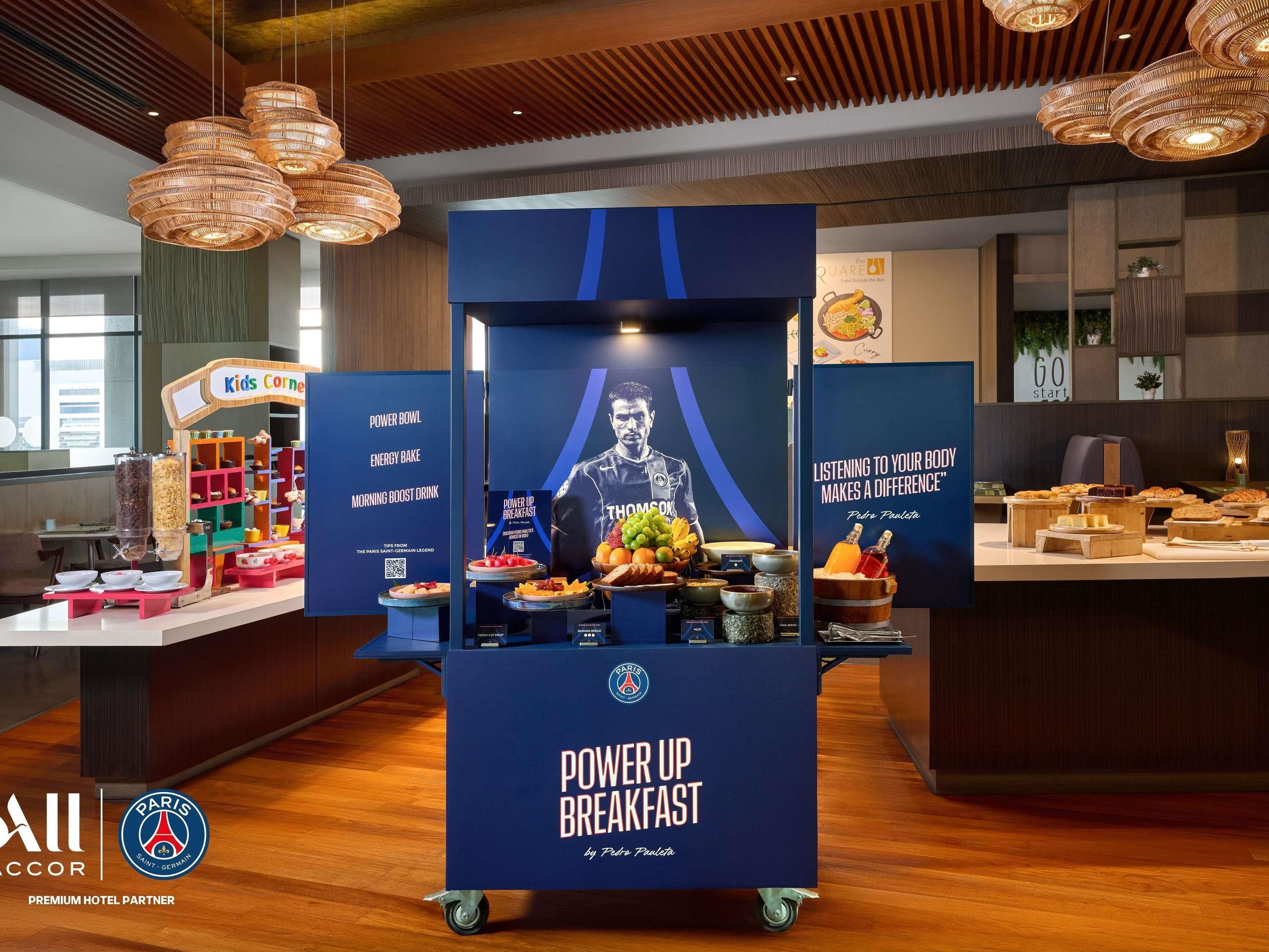Een ontbijtruimte met een 'Power Up Breakfast'-station, met diverse etenswaren en een poster met een sportief thema.