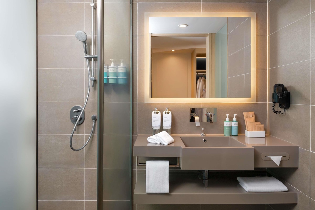 A modern bathroom with a glass-enclosed shower, a large mirror, a sink with toiletries, and a telephone.