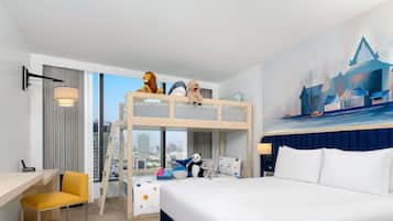 A hotel room with a bunk bed, a desk, a chair, a bed with white bedding, a city view, and a painting on the wall.