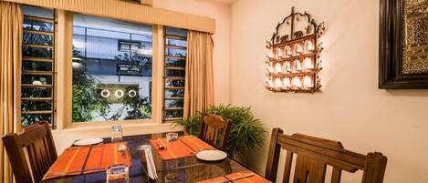 Un comedor con una mesa lista para cuatro, ventanas amplias con cortinas, una lámpara fijada a la pared y una planta en maceta.