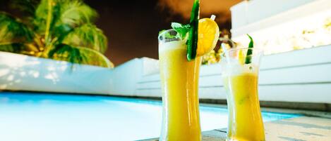 Two tall glasses of a yellow beverage with lime and mint garnish, placed on a wooden surface near a pool with palm trees in the background.