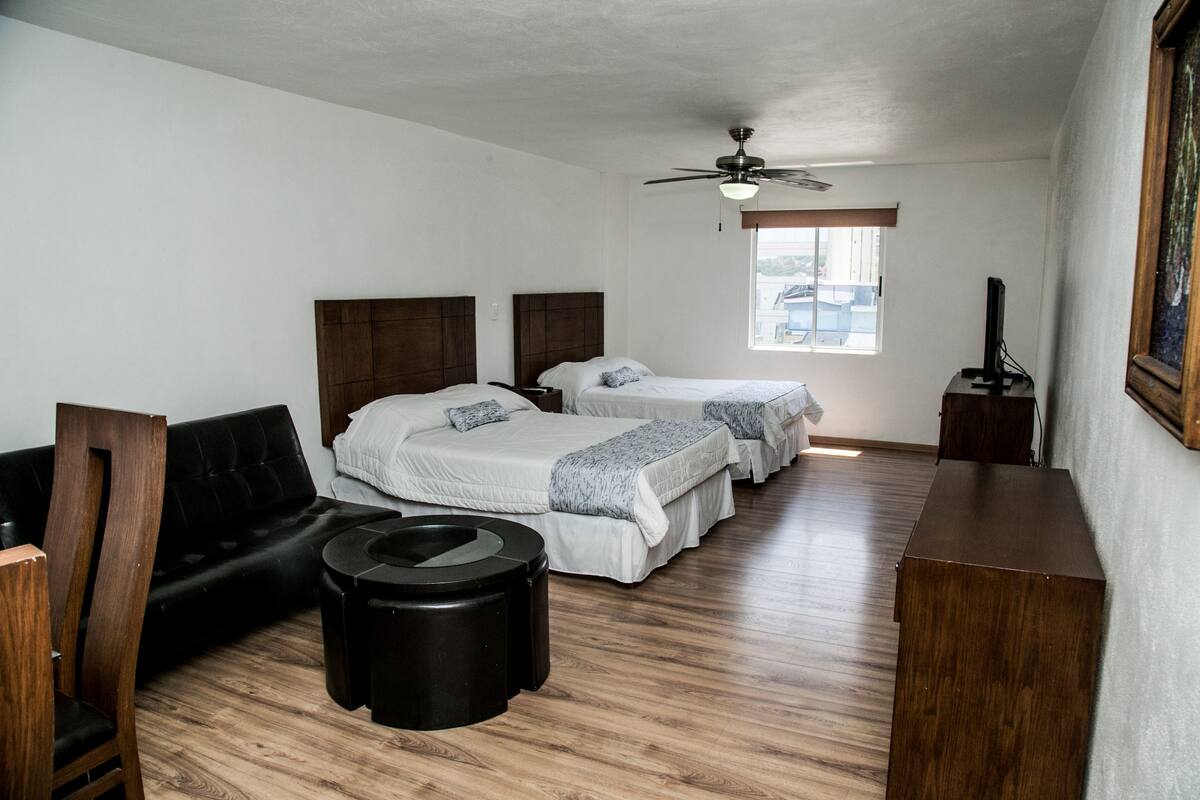 A hotel room with two beds, a black sofa, a wooden chair, a small table, and a ceiling fan.