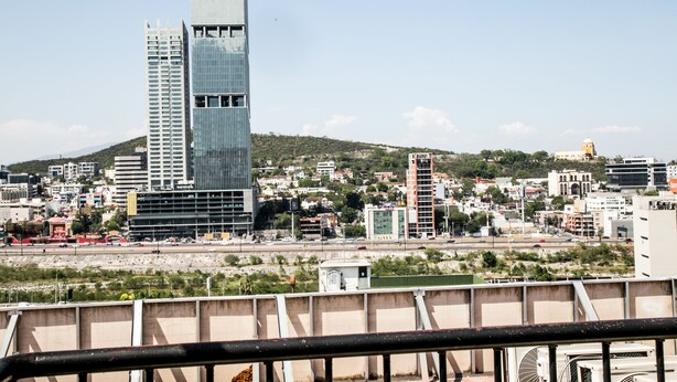 A tall glass skyscraper with a modern design, surrounded by a cityscape with various buildings and a hilly background. - Suites DIOH (Monterrey)