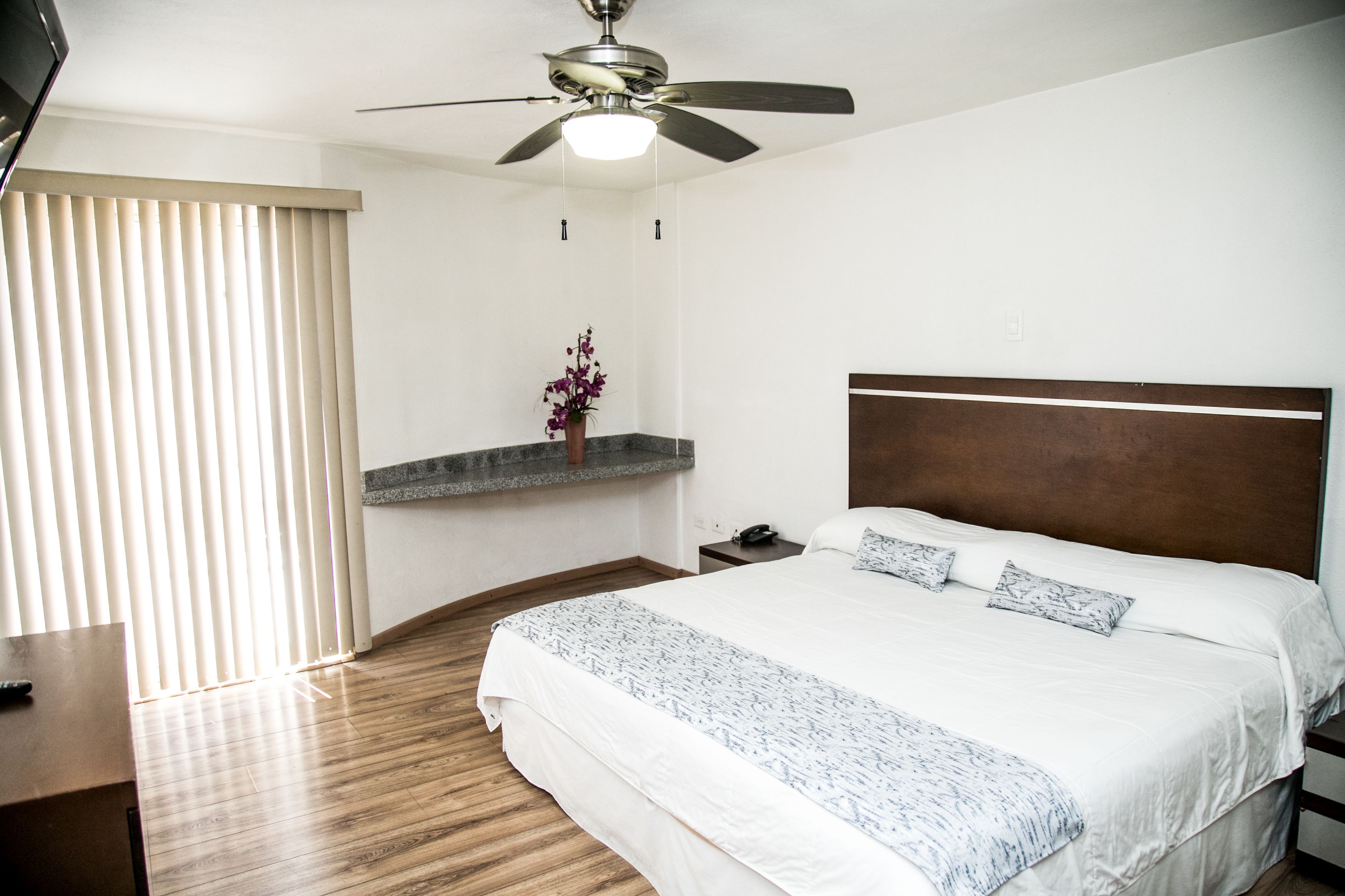 A bedroom with a bed, a nightstand, a TV, a ceiling fan, and a window with blinds.