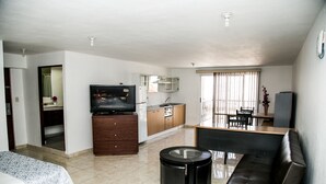 A modern living room with a sofa, a coffee table, a TV, and a kitchen area with cabinets and a refrigerator. - Suites DIOH (Monterrey)