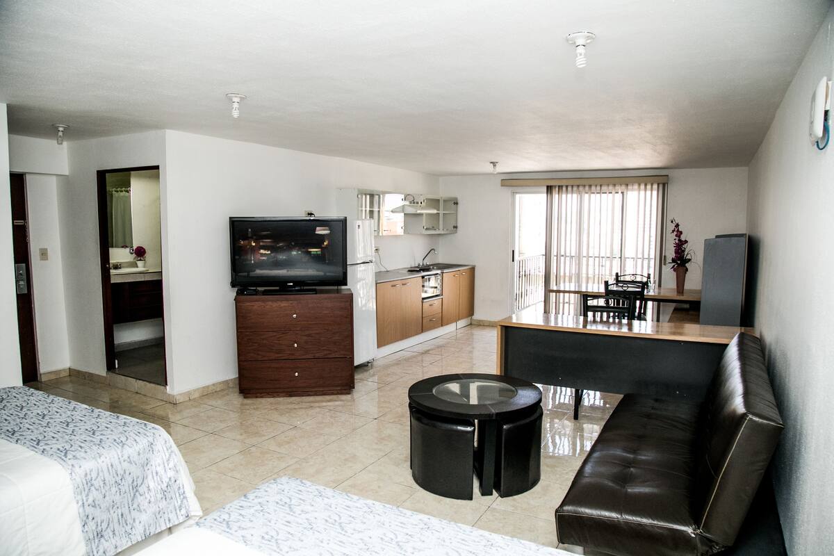 A modern living room with a sofa, a coffee table, a TV, and a kitchen area with cabinets and a refrigerator.
