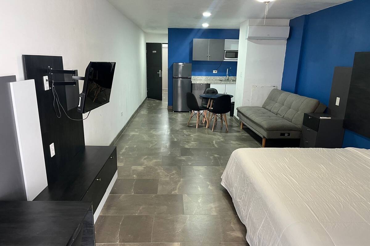 A modern hotel room with a bed, a sofa, a desk with chairs, a kitchenette, and a TV mounted on the wall.