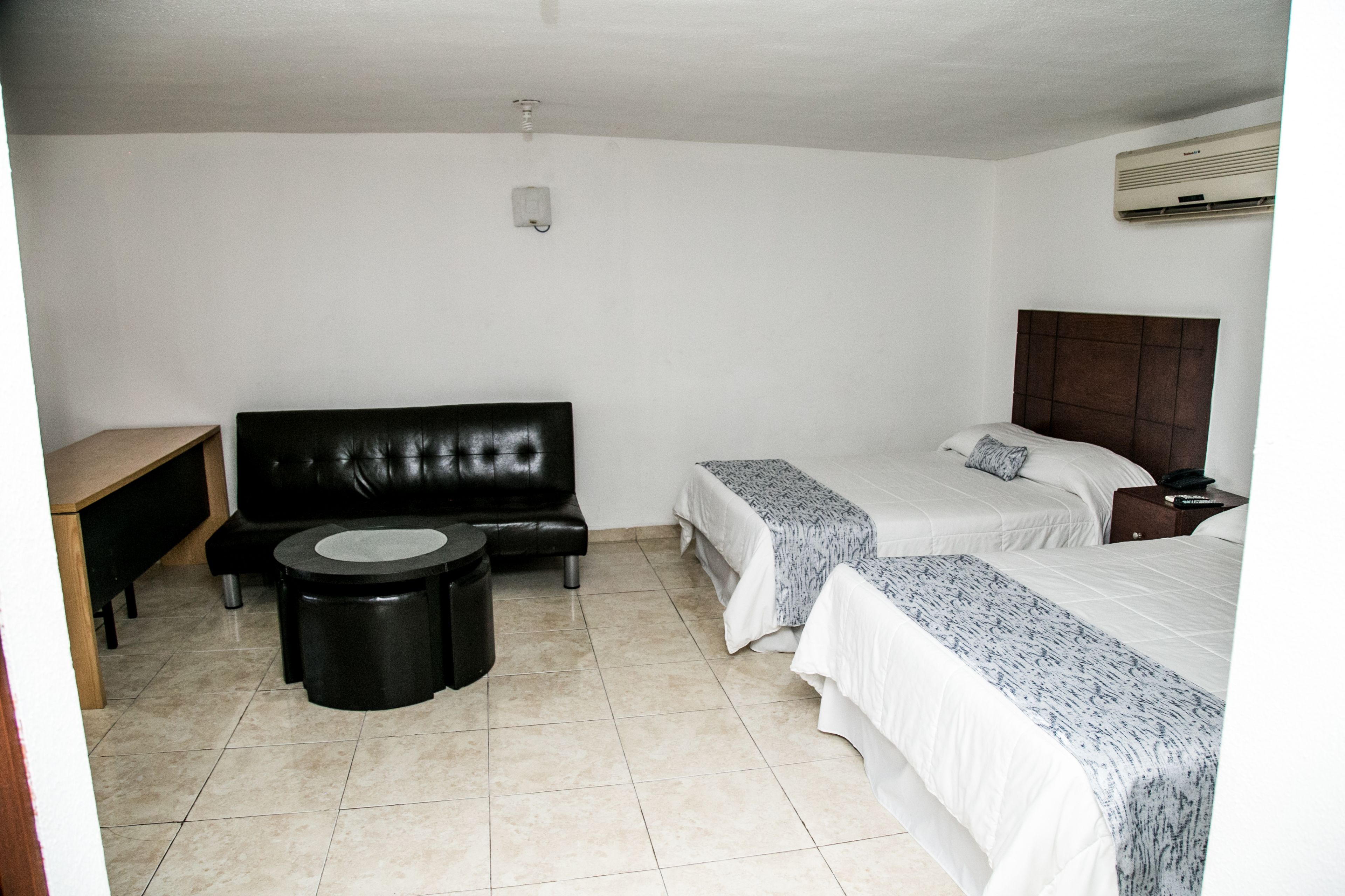 A hotel room with two beds, a black leather sofa, a small round table, and a bedside table with a lamp.