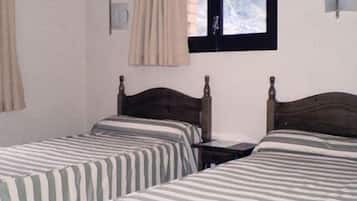 Quarto com duas camas individuais, uma janela com cortinas e um ponto de luz fixado na parede.