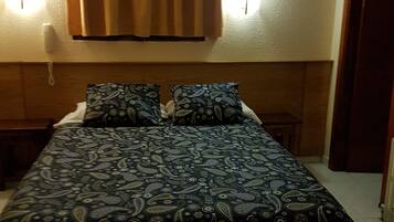 Un dormitorio con cama, dos mesitas de noche, un teléfono en la pared y una ventana con cortinas.