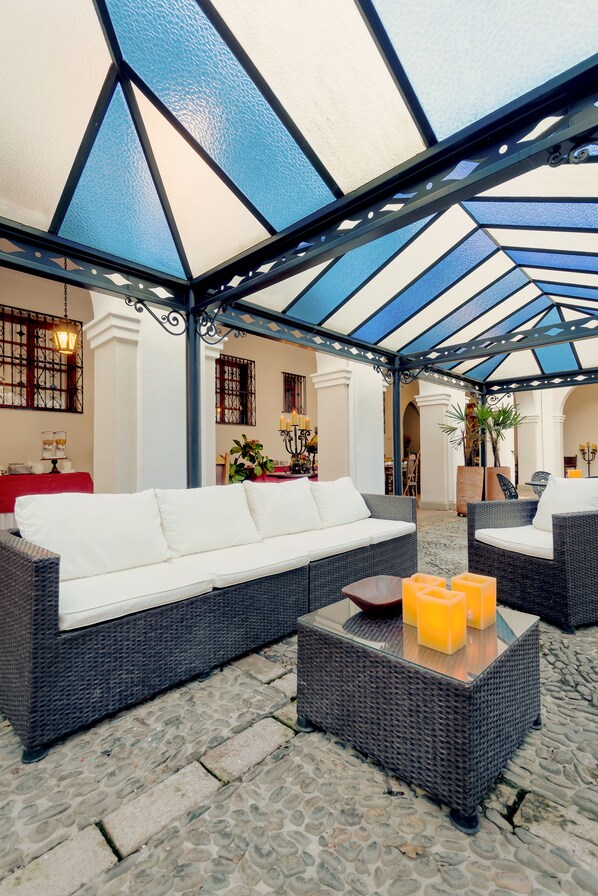 Un espacio de estar al aire libre con techo, mobiliario de mimbre y una mesa con superficie de vidrio.
