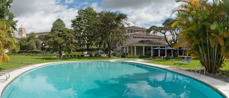 A swimming pool with a clear blue water, surrounded by lush greenery and a building with a dome in the background.