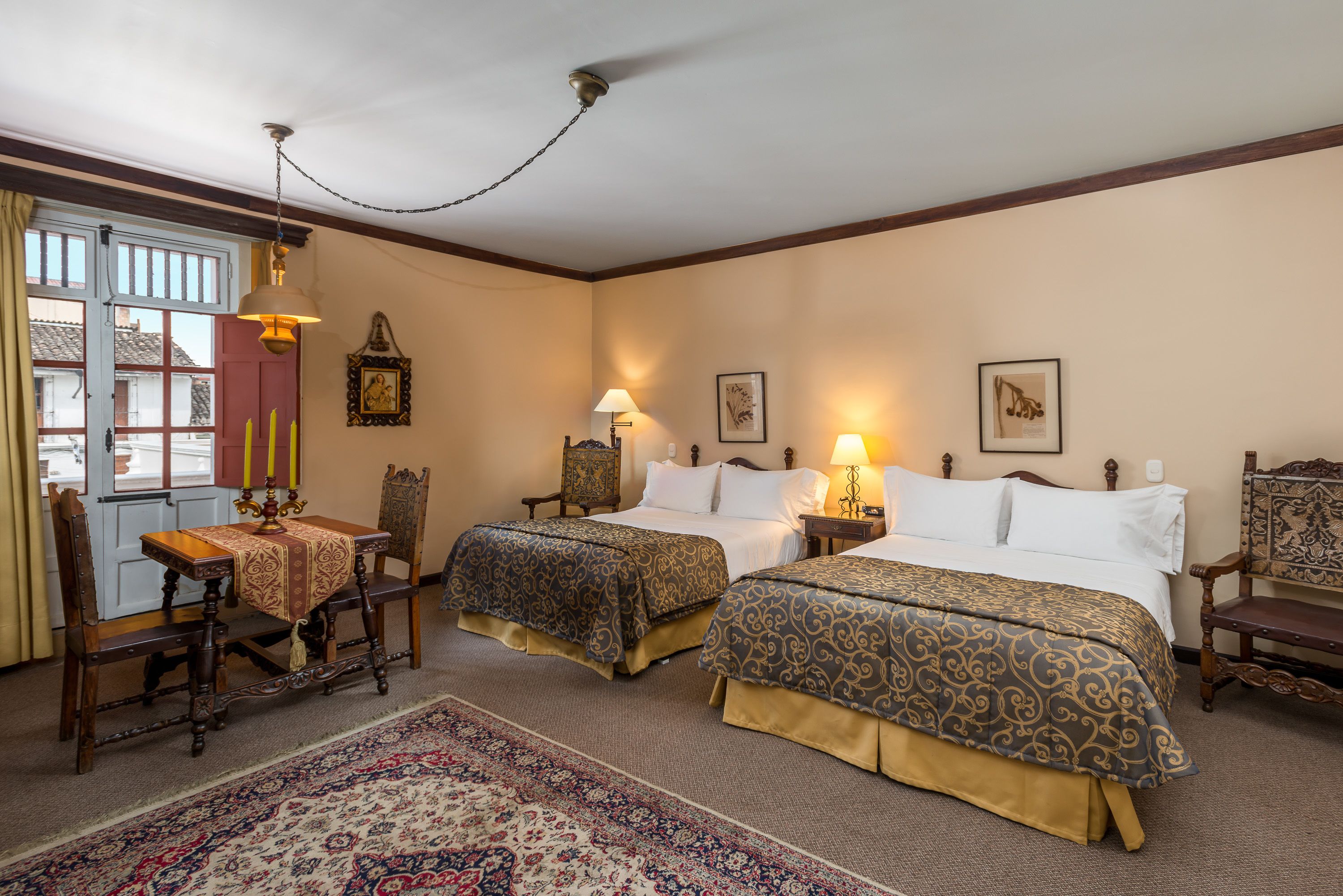 A hotel room with two beds, a dining table, chairs, a chandelier, and a large window with curtains.