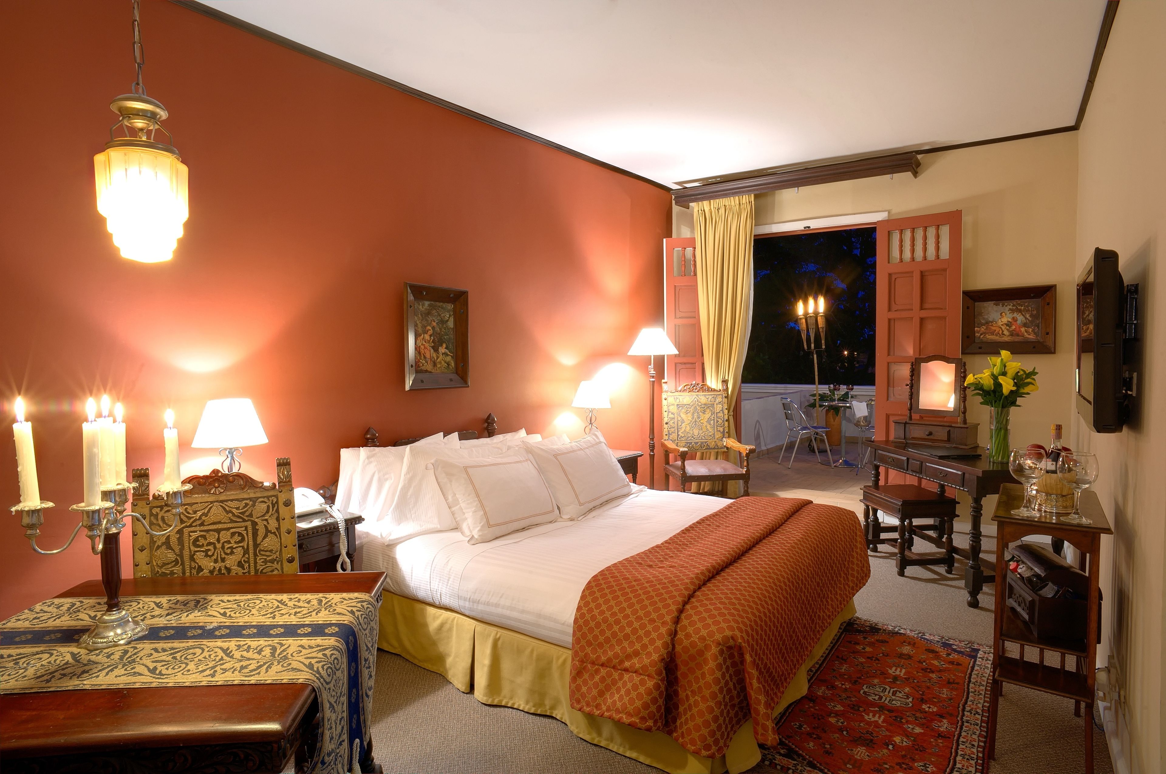 A hotel room with a bed, bedside lamps, a desk with a chair, a small table with a vase, and a view of the outside.