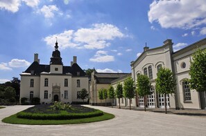 A large, historic building with a prominent tower and symmetrical architecture, surrounded by a well-maintained garden and a gravel pathway.
