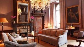 Een klassieke woonkamer met een open haard, een kroonluchter en een groot portret aan de muur.