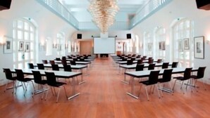 Een congreszaal met rijen tafels en stoelen, een grote kroonluchter en een whiteboard aan de voorzijde.