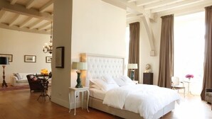 Een slaapkamer met een groot bed, een wit hoofdeinde, een nachtkastje met een lamp, een stoel en een tafeltje met een bloevas.
