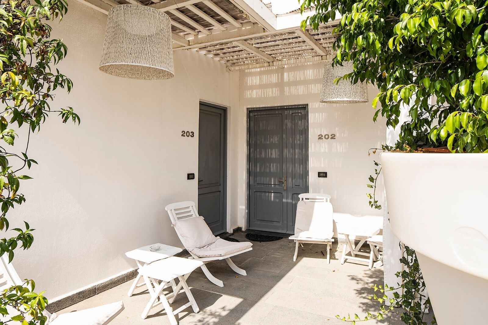 A sunlit outdoor seating area with two lounge chairs and a small table, adjacent to a building with two numbered doors, 203 and 202.