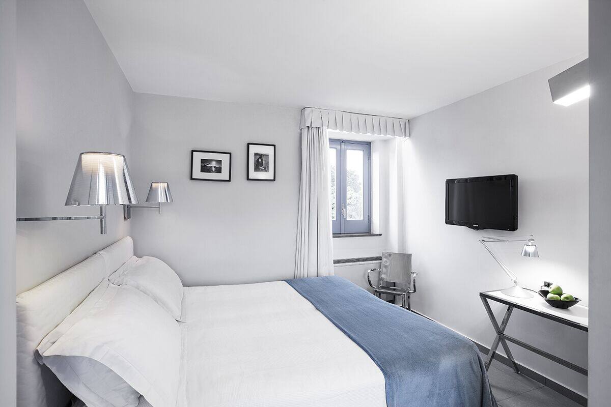 A modern hotel room with a bed, a desk with a lamp, a TV, and a window with curtains.