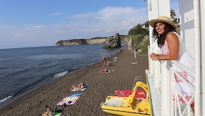 A beach scene with a person leaning on a white structure, a sandy beach, and a rocky cliff in the background. - La Suite Boutique Hotel (Procida)