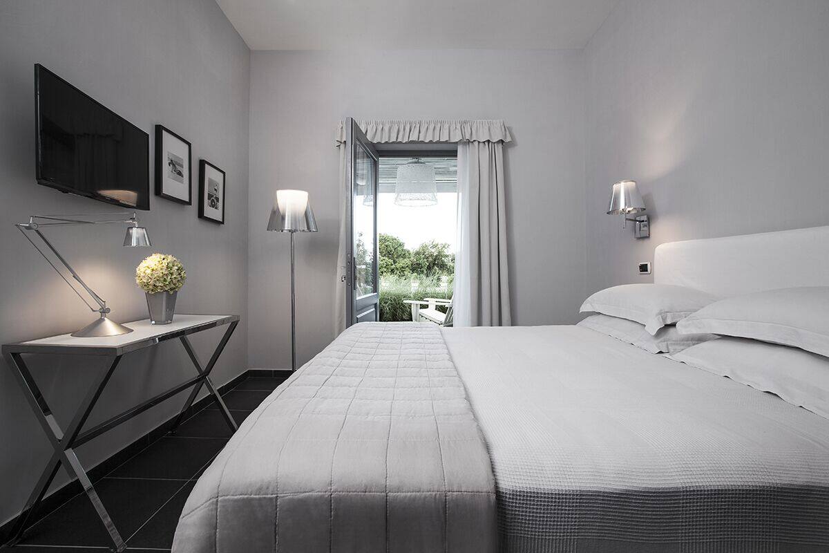 A modern hotel room with a large bed, a desk with a lamp, a potted plant, and a view of greenery outside.