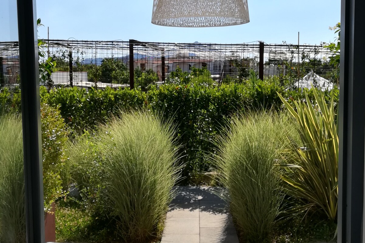 Eine Terrasse mit weißen Liegestühlen, einer hängenden Leuchte und Blick auf einen Garten sowie entfernte Gebäude.