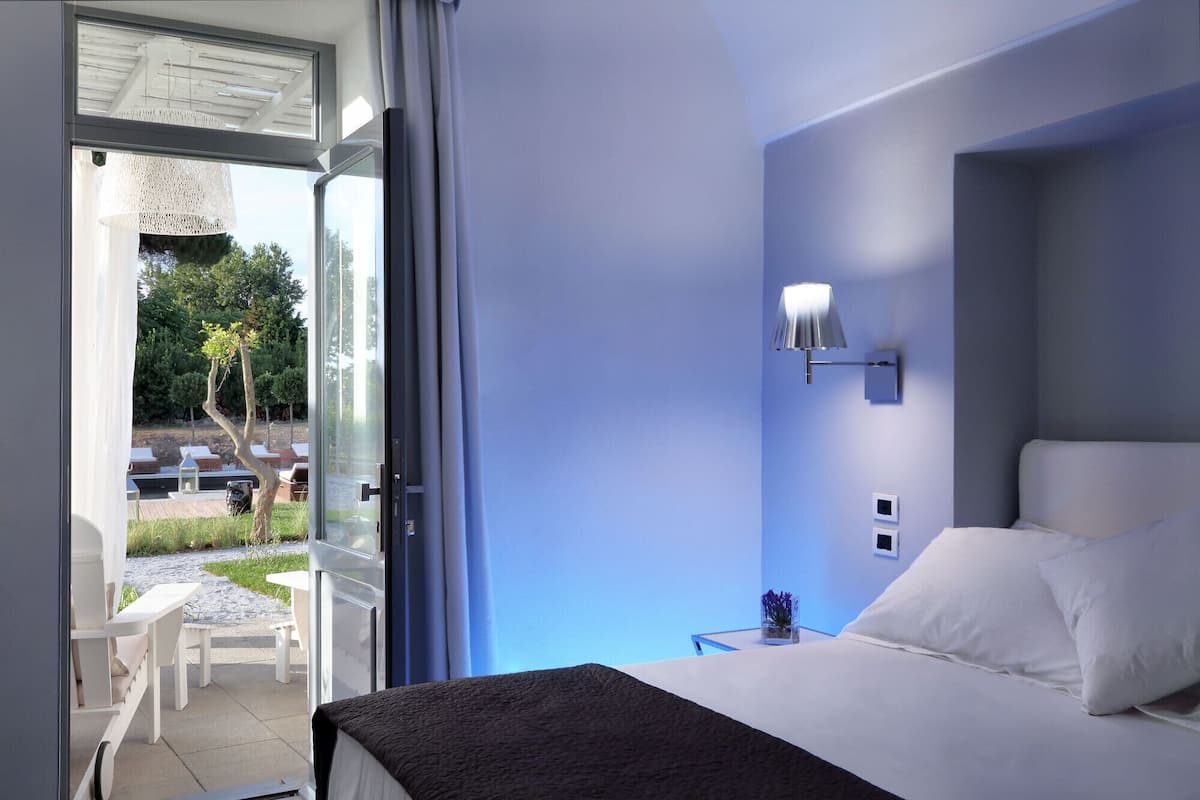 A hotel room with a bed, a nightstand, a wall-mounted lamp, and a view of the outdoor patio area.