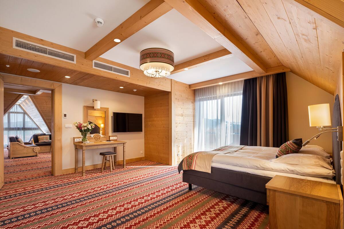 Une chambre d’hôtel avec un plafond en pente, des poutres apparentes, un grand lit, un bureau et une vue sur l’extérieur.