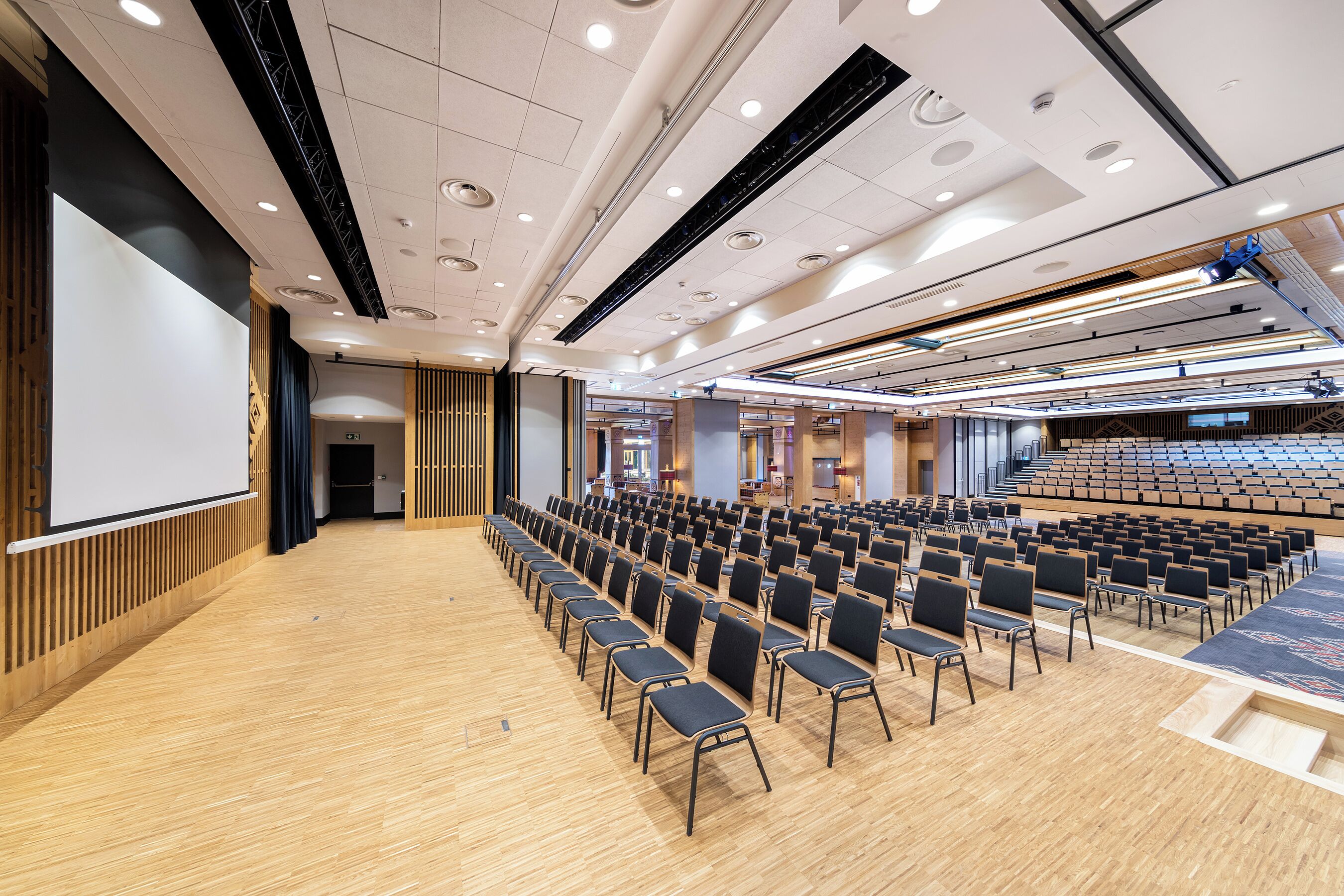 Un salón de conferencias con filas de sillas, una pantalla grande y una zona escenario.