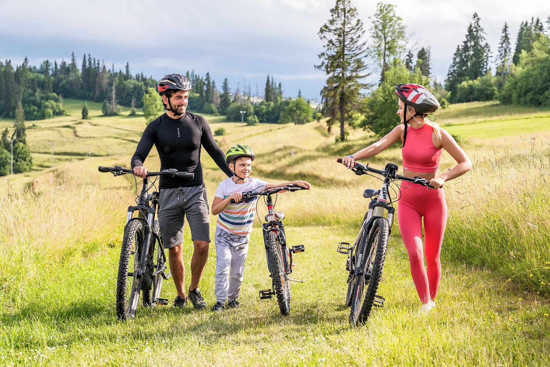 A family with three bicycles in a grassy field with trees in the background.