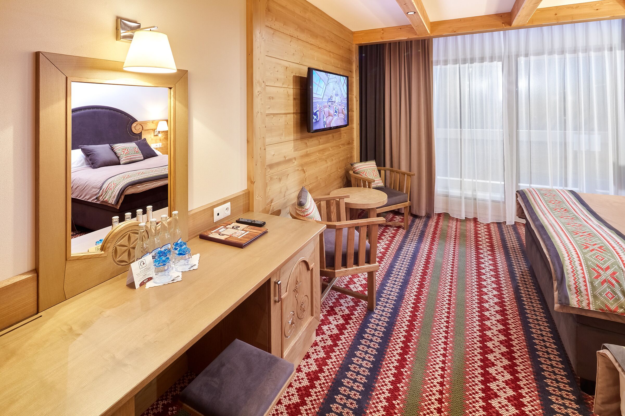 Een hotelkamer met een bed met houten hoofdeinde, een aan de muur bevestigde televisie, een houten bureau met stoel, en een kamerbreed tapijt met een patroon.