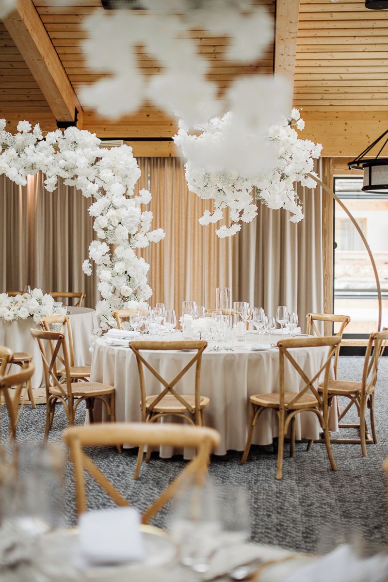 Decoración para una recepción de boda con mesas redondas, arreglos florales blancos y sillas de madera.