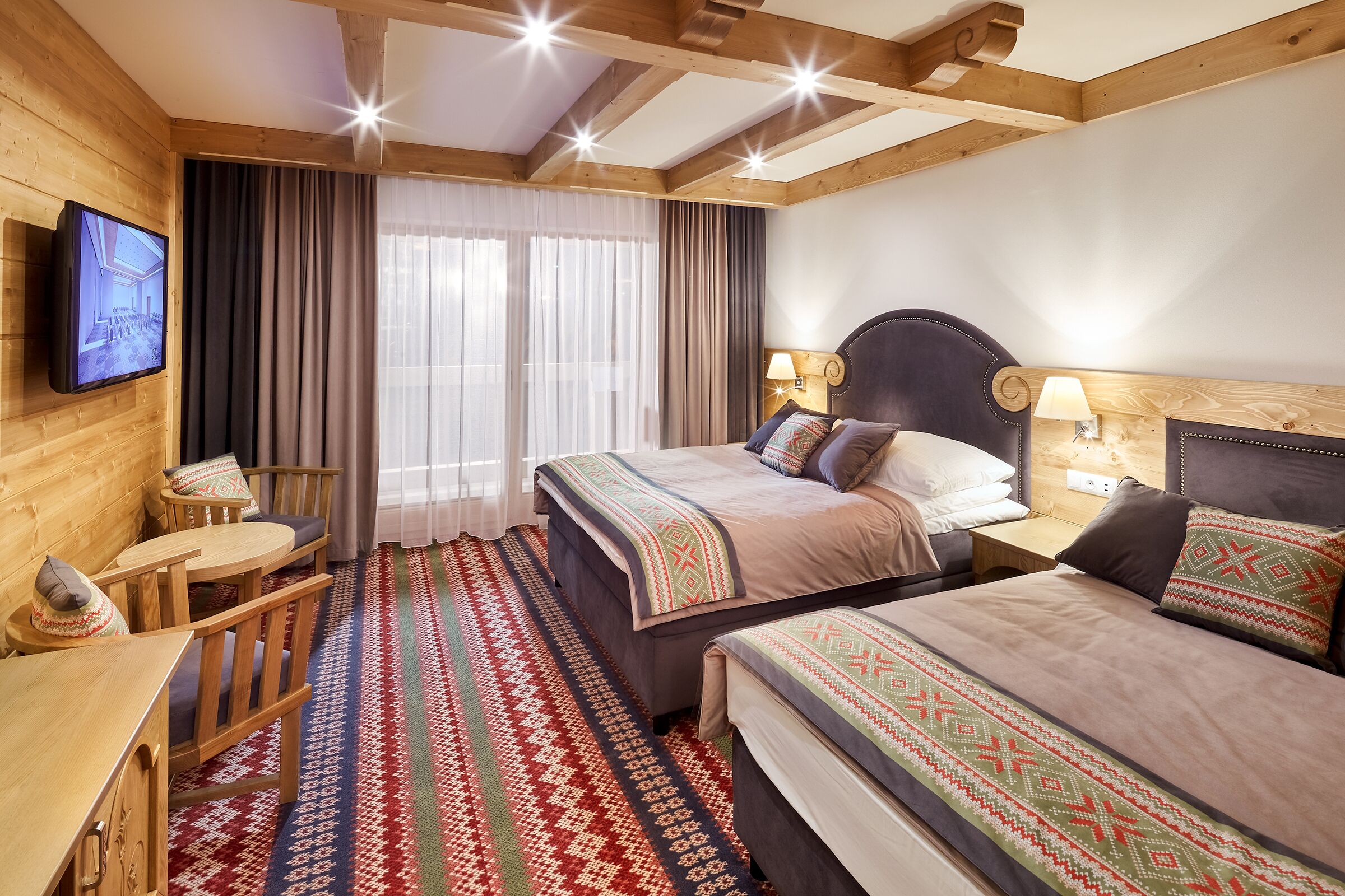 Habitación de hotel con dos camas, cabecero de madera, televisor montado en la pared, una mesita pequeña y una alfombra estampada.
