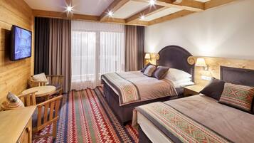 Habitación de hotel con dos camas, cabecero de madera, televisor montado en la pared, una mesita pequeña y una alfombra estampada.