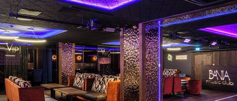 Un área de lounge moderna con asientos y un escenario, con paneles de techo iluminados y un letrero que dice 'Bania Music Club'.