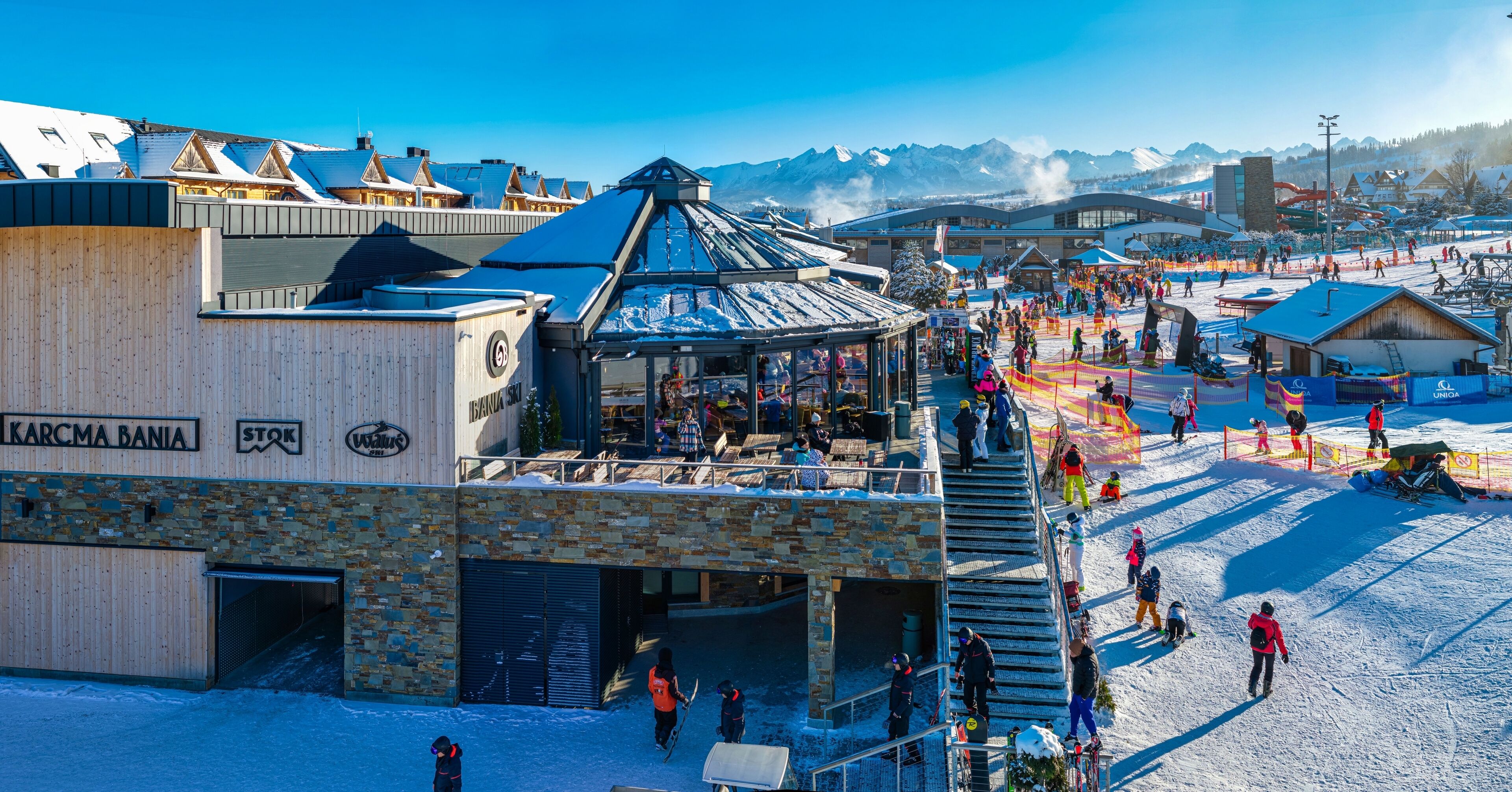 A ski resort with a wooden building labeled "KARCMA BANIA" and "STOK," featuring a ski lift, a covered area with people, and a snowy landscape.