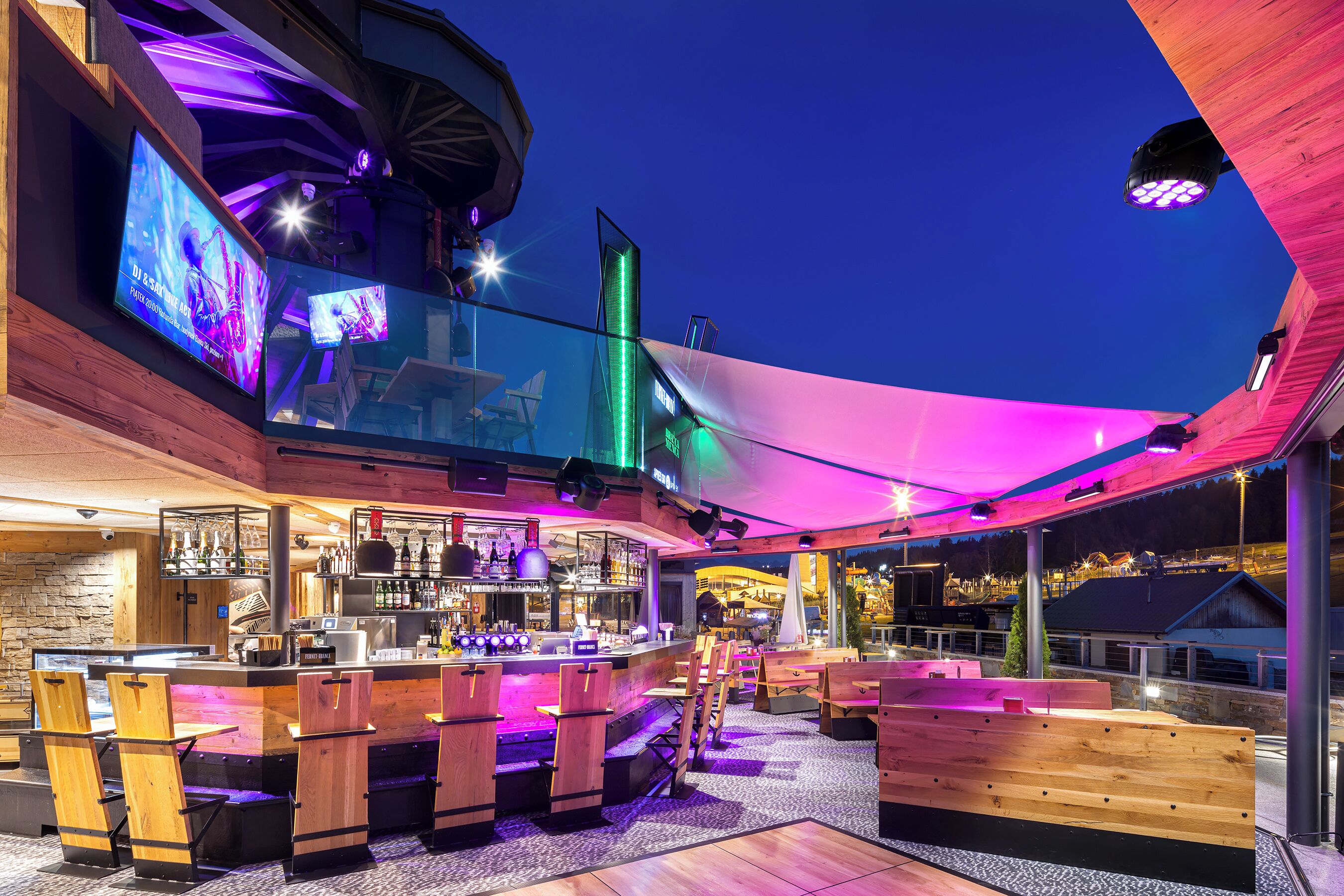 Un área de bar al aire libre con mobiliario de madera, una barra y una pantalla grande que muestra un video.