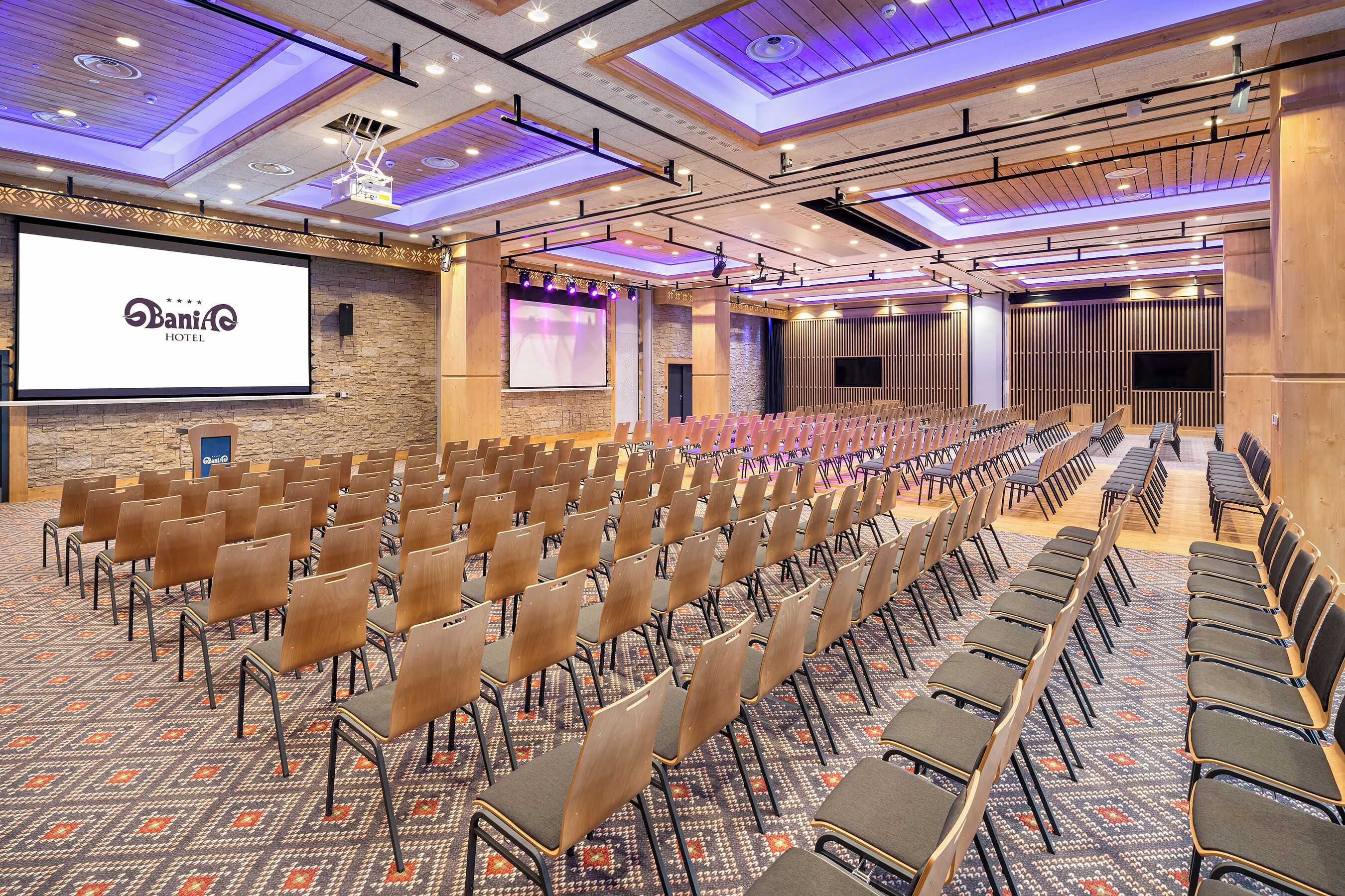 Un salón de conferencias con filas de sillas, una pantalla grande y un proyector.
