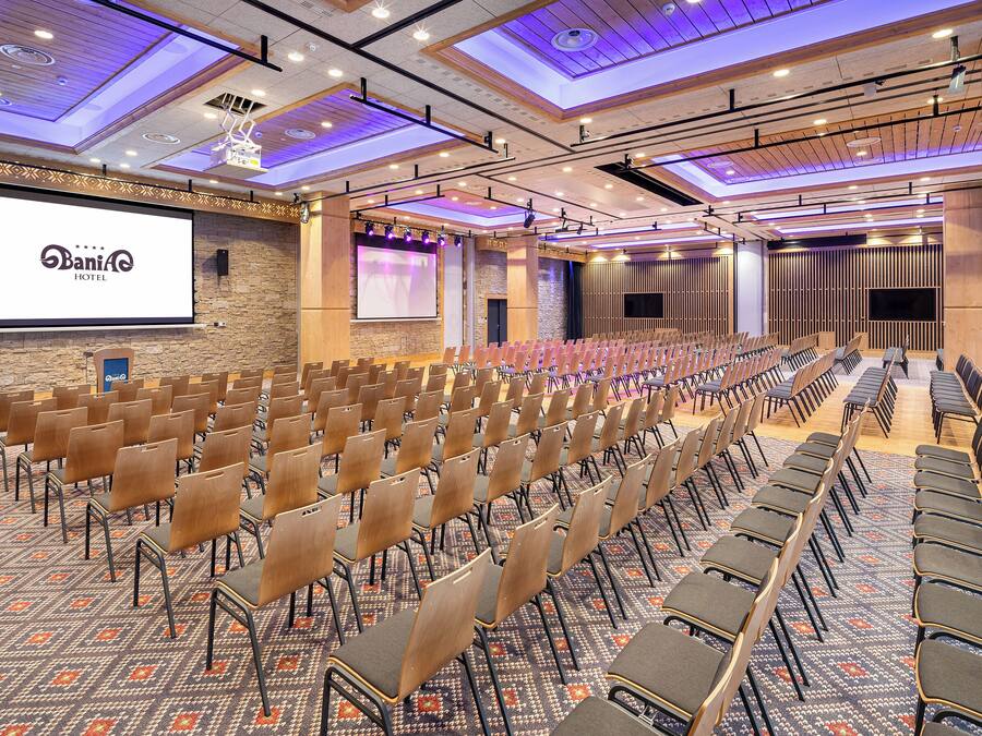 Un salón de conferencias con filas de sillas, una pantalla grande y un proyector.