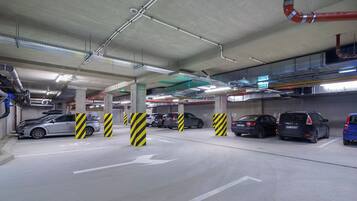 Un estacionamiento subterráneo de varios niveles con varios coches aparcados, pilares de concreto con franjas amarillas y negras, y flechas direccionales en el suelo.