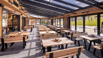 Un espacio de comedor al aire libre con mesas y bancos de madera, grandes ventanales y vistas al parque.