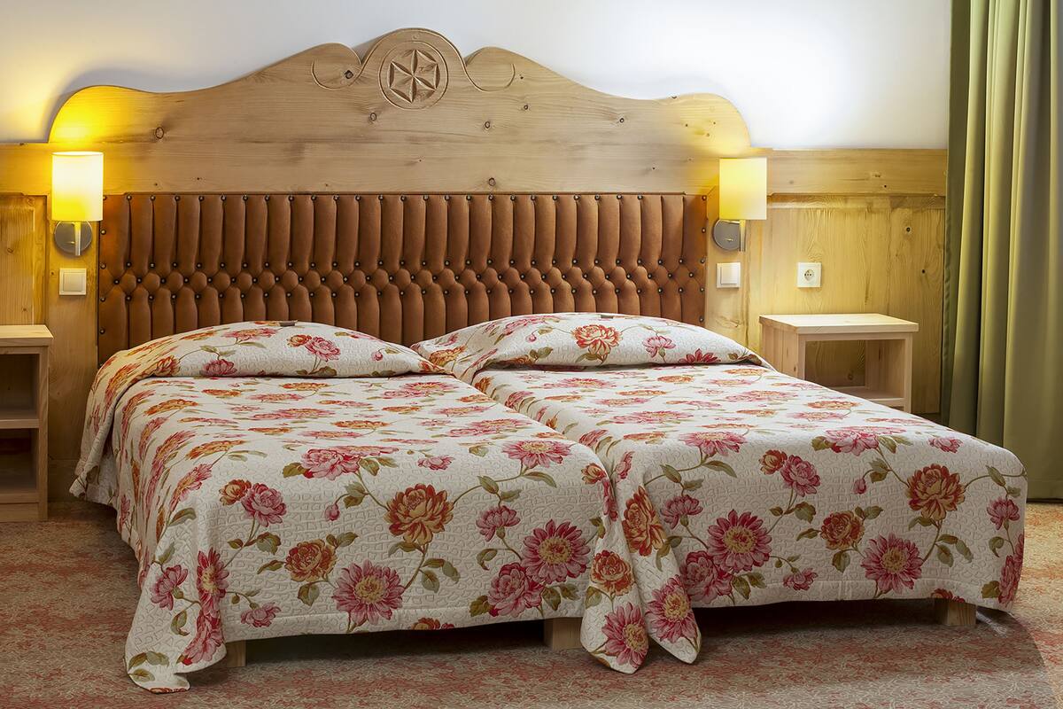 Un lit double avec une literie à motifs floraux, une tête de lit en bois, deux tables de chevet avec des lampes et une petite table de nuit en bois.