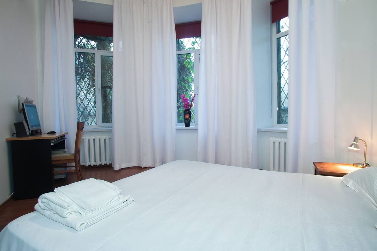 A neatly made bed in a room with a desk, chair, and a window with curtains.
