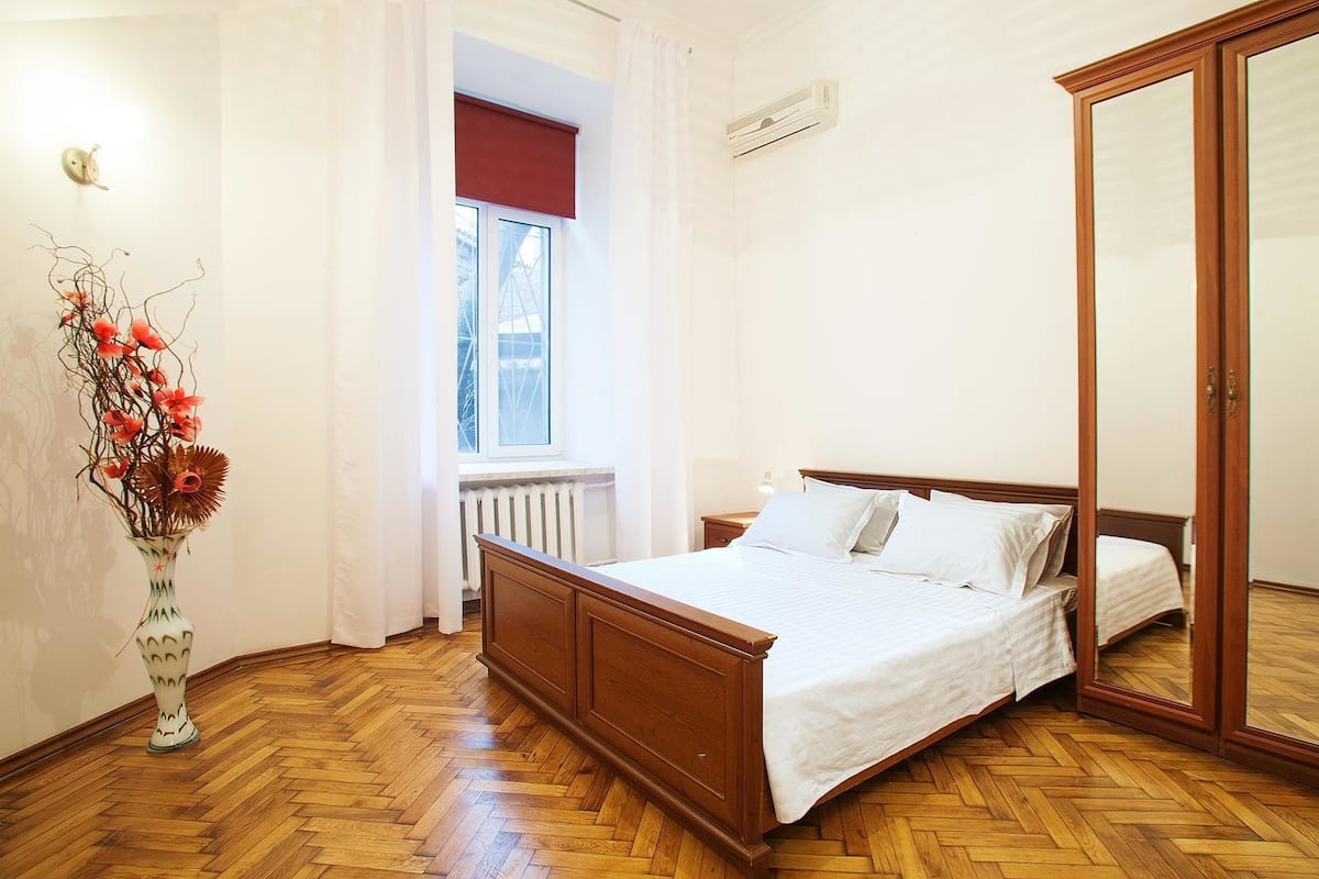 A bedroom with a wooden bed, white bedding, a radiator, a window with a red blind, and a wooden wardrobe.