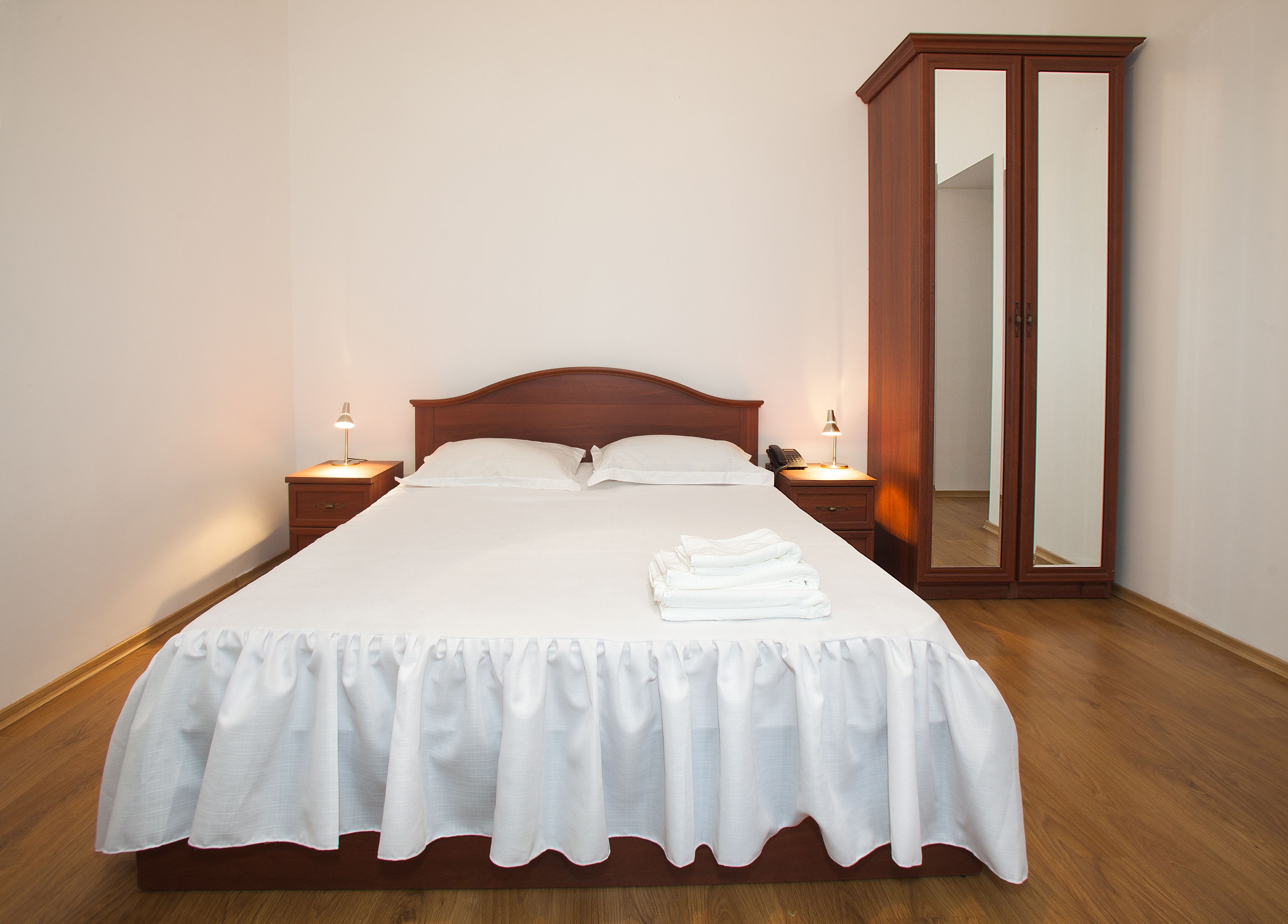 Een opgemaakt bed met een witte spreideken en kussens, twee nachtlampjes, een houten nachtkastje en een grote spiegel aan de muur.