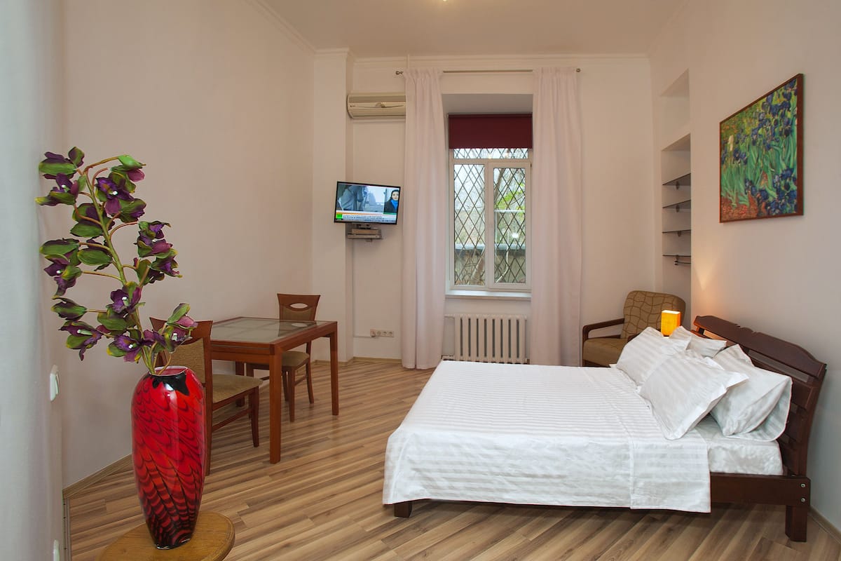 A hotel room with a bed, a TV, a dining table, and a vase of flowers.